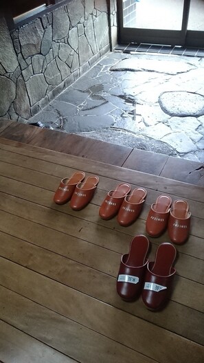 A set of brown leather slippers with the brand name 'KOMENKO' on a wooden floor near a stone wall and a glass door.