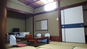 A traditional Japanese room with tatami flooring, a low table, chairs, a television, and a wardrobe. - Komecho Ryokan (Imabari)