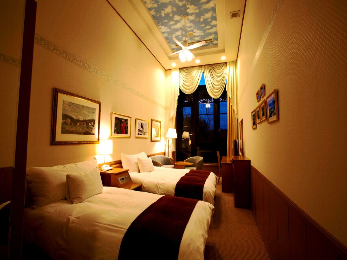A hotel room with two beds, a ceiling fan, a nightstand, a desk, a chair, and a window with curtains.