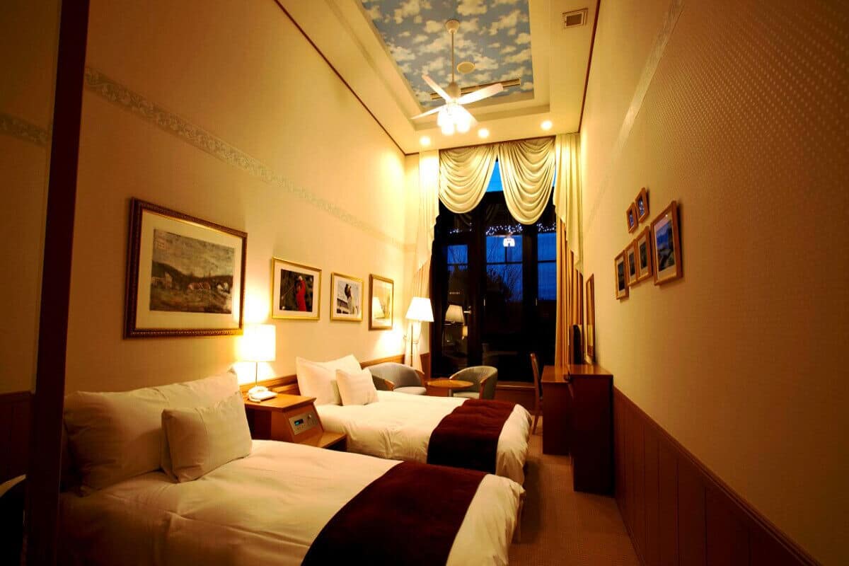 A hotel room with two beds, a ceiling fan, a nightstand, a desk, a chair, and a window with curtains.