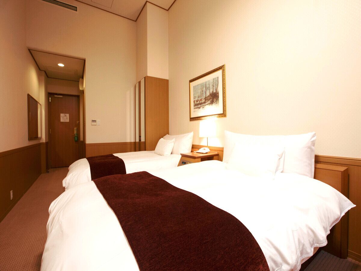 A hotel room with two beds, a nightstand, a lamp, a painting on the wall, and a door leading to another room.
