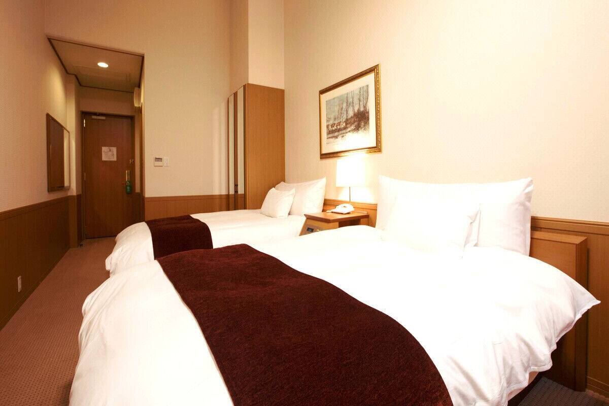 A hotel room with two beds, a nightstand, a lamp, a painting on the wall, and a door leading to another room.