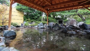 A traditional Japanese onsen with a wooden pavilion, a stone bath surrounded by rocks, and a view of greenery.