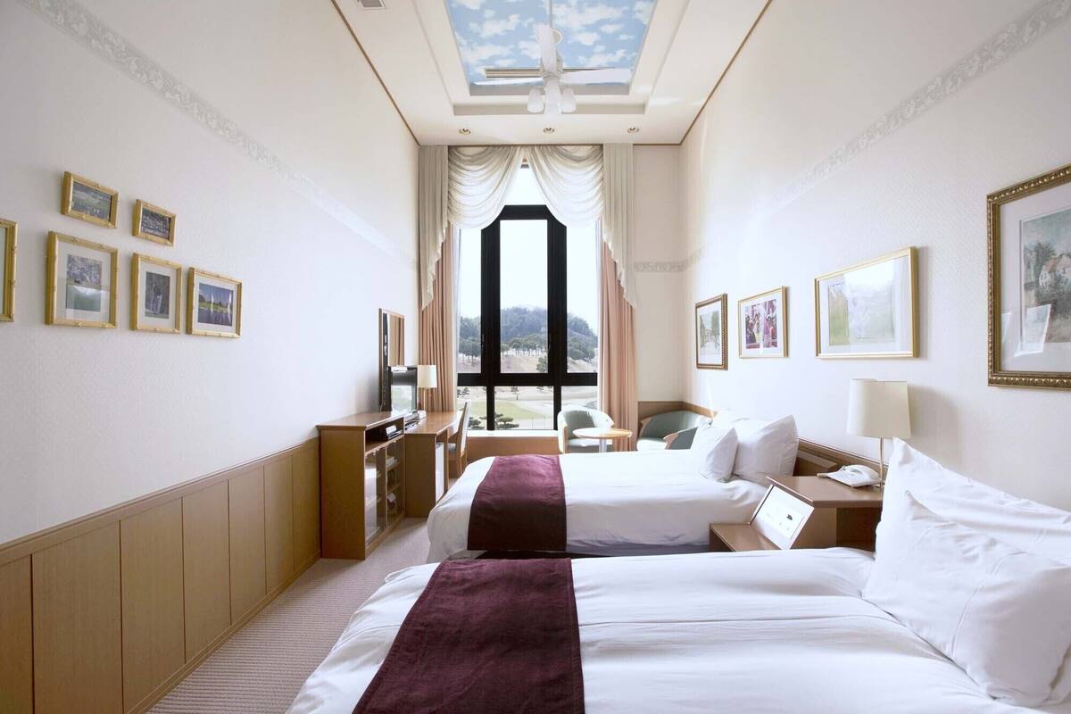 A hotel room with two beds, a desk with a chair, a TV, and a large window with a view of trees.