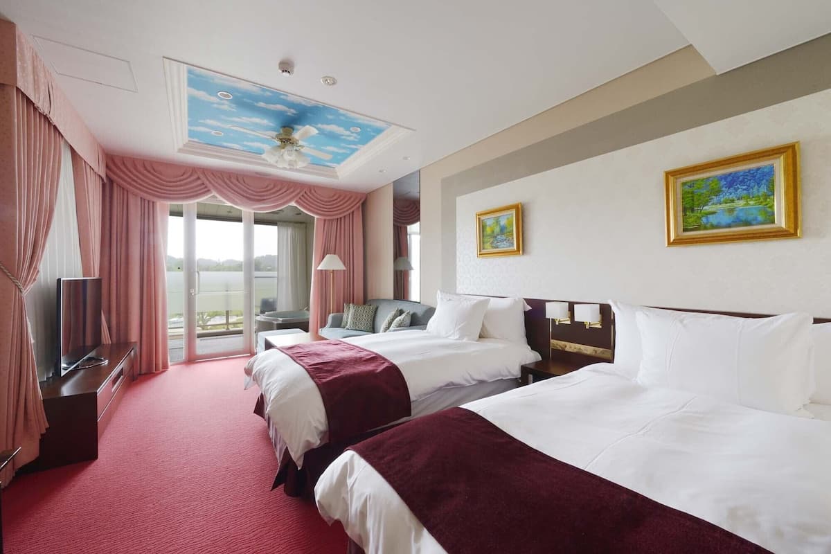 A hotel room with two beds, a TV, a desk, and a balcony with curtains.