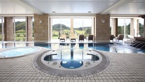 A large indoor swimming pool with a hot tub, lounge chairs, and a view of a green landscape through large windows.