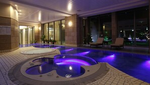 A heated outdoor swimming pool area with two hot tubs, lounge chairs, and a view of the cityscape at night.