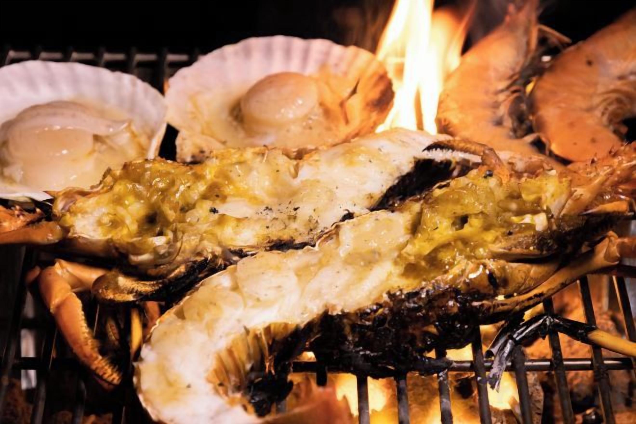 Grilled seafood, including lobster and clams, being cooked over an open flame.