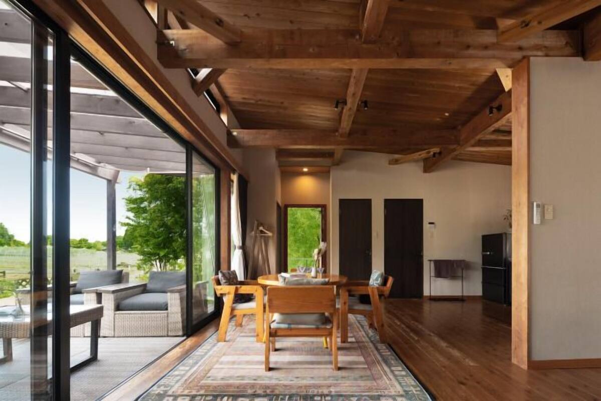 A modern interior with a wooden ceiling, a dining area with a table and chairs, and large windows offering a view of the outdoors.