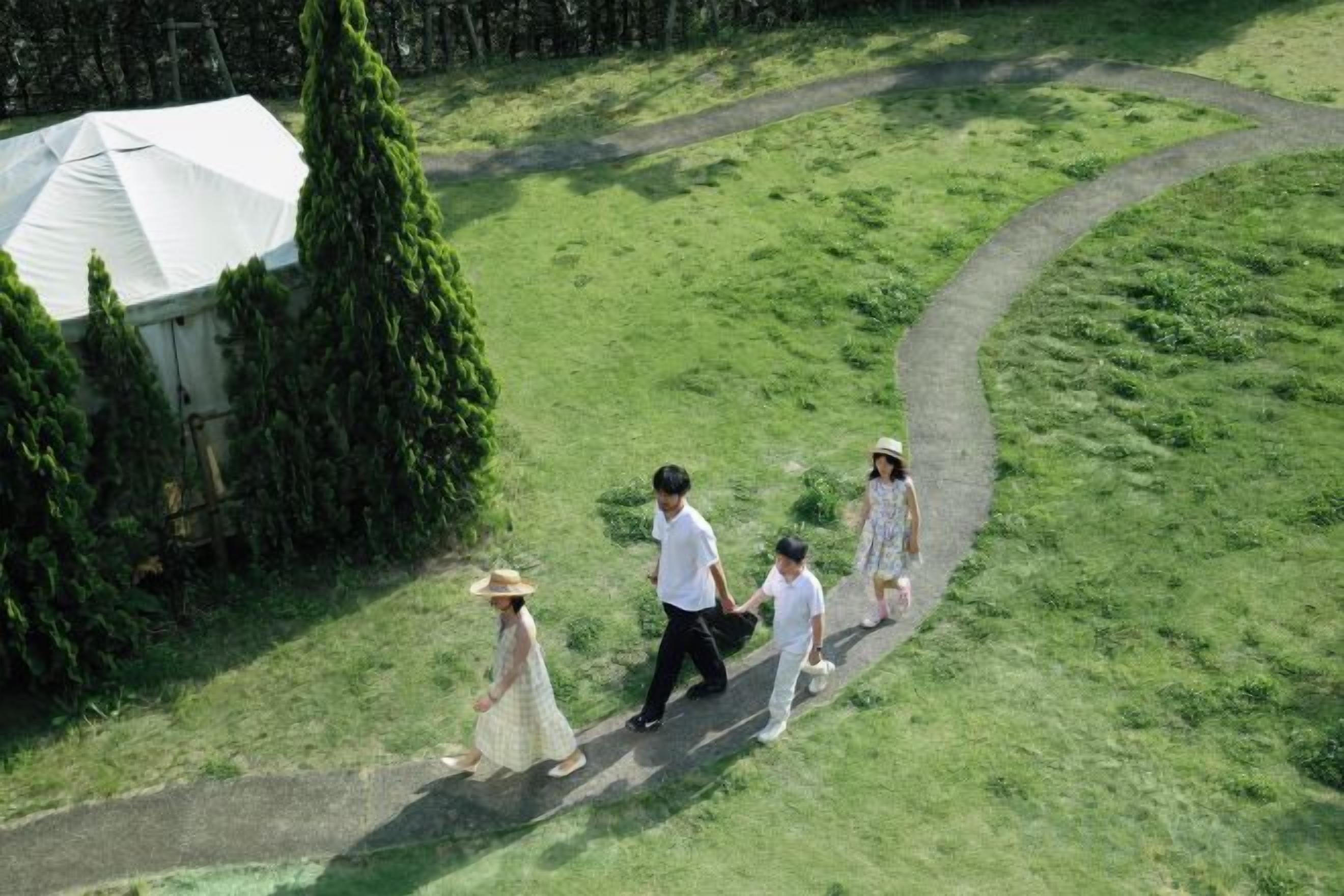 A group of people walking on a grassy path with a tent and trees in the background.