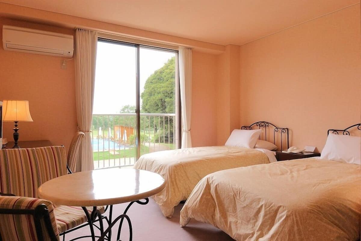 Une chambre d’hôtel avec deux lits, une table ronde, une chaise, une lampe et une vue sur un balcon agrémenté de verdure.