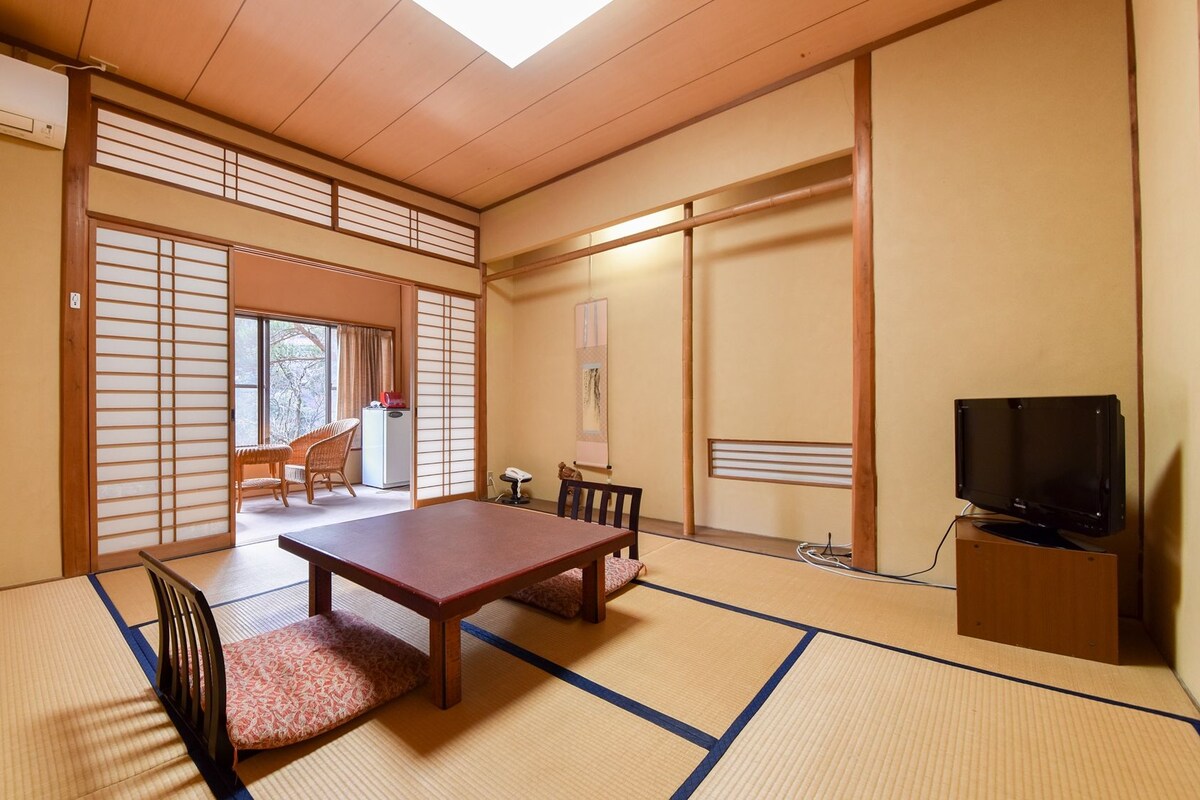 Una habitación tradicional japonesa con suelo de tatami, una mesa baja, sillas, televisor y puertas correderas.