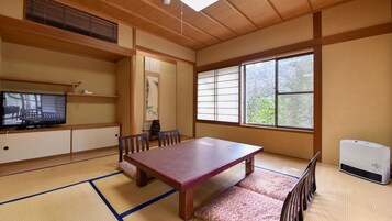 Una habitación tradicional japonesa con suelo de tatami, una mesa baja, sillas, un televisor y un gran ventanal con vistas a árboles.