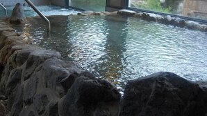 A hot spring area with a stone wall, a metal ladder, and a view of the outdoors through a large window. - Yamae Onsen Hotaru (Yamae)