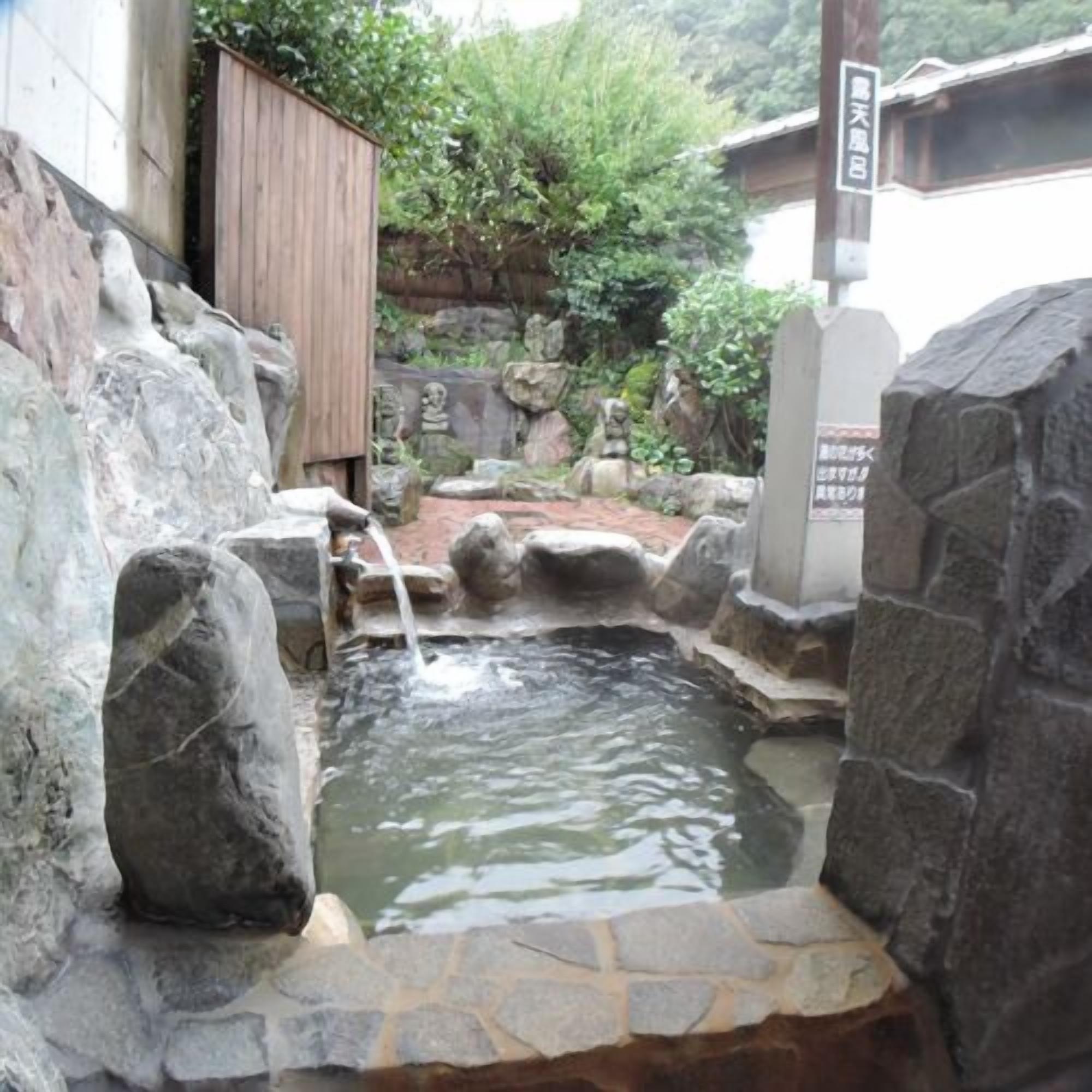 Um jardim japonês tradicional com lago de pedra, estruturas de madeira e uma placa em japonês.