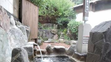 Um jardim japonês tradicional com lago de pedra, estruturas de madeira e uma placa em japonês.