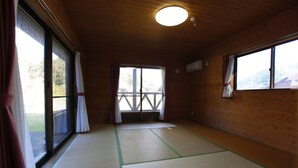 Una habitación con esteras de tatami, paredes de madera, ventanas amplias con cortinas y vista a una terraza.