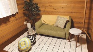 Una habitación acogedora con suelo de madera, un sofá verde con una manta amarilla, una mesita redonda, una planta en maceta y un reloj de pared.