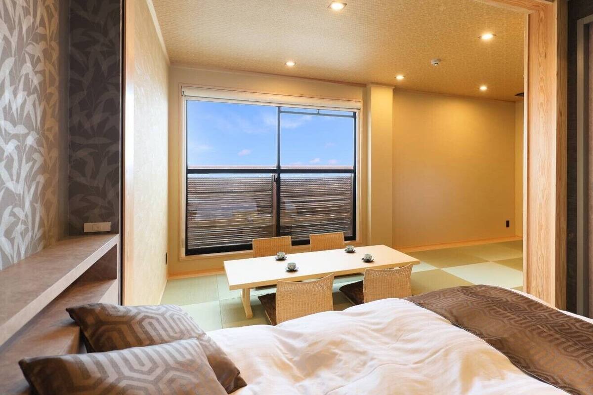 Une pièce comprenant un lit, une table à manger avec des chaises, une fenêtre avec des stores et une porte.
