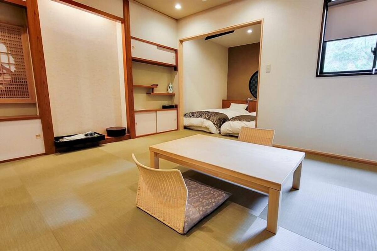 Une pièce de style japonais traditionnel, avec un sol recouvert de tatamis, une petite table basse, une chaise et un lit équipé d’un futon.