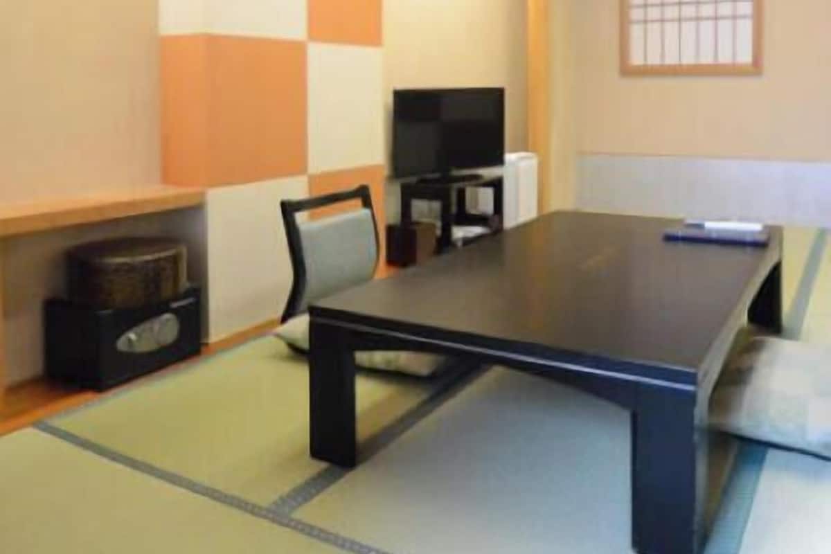 Una habitación con suelo de tatami, una mesa baja, una silla, un televisor y un estante con un altavoz.