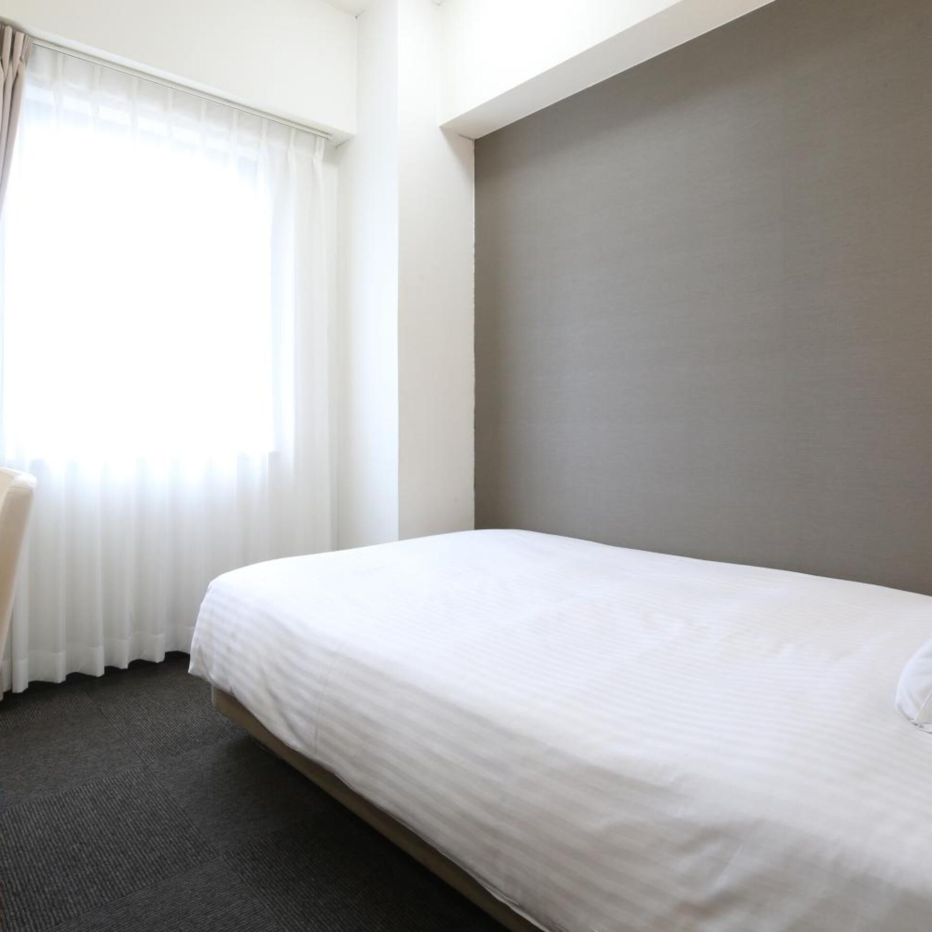 A hotel room with a single bed, a chair, a window with curtains, and a plain wall.