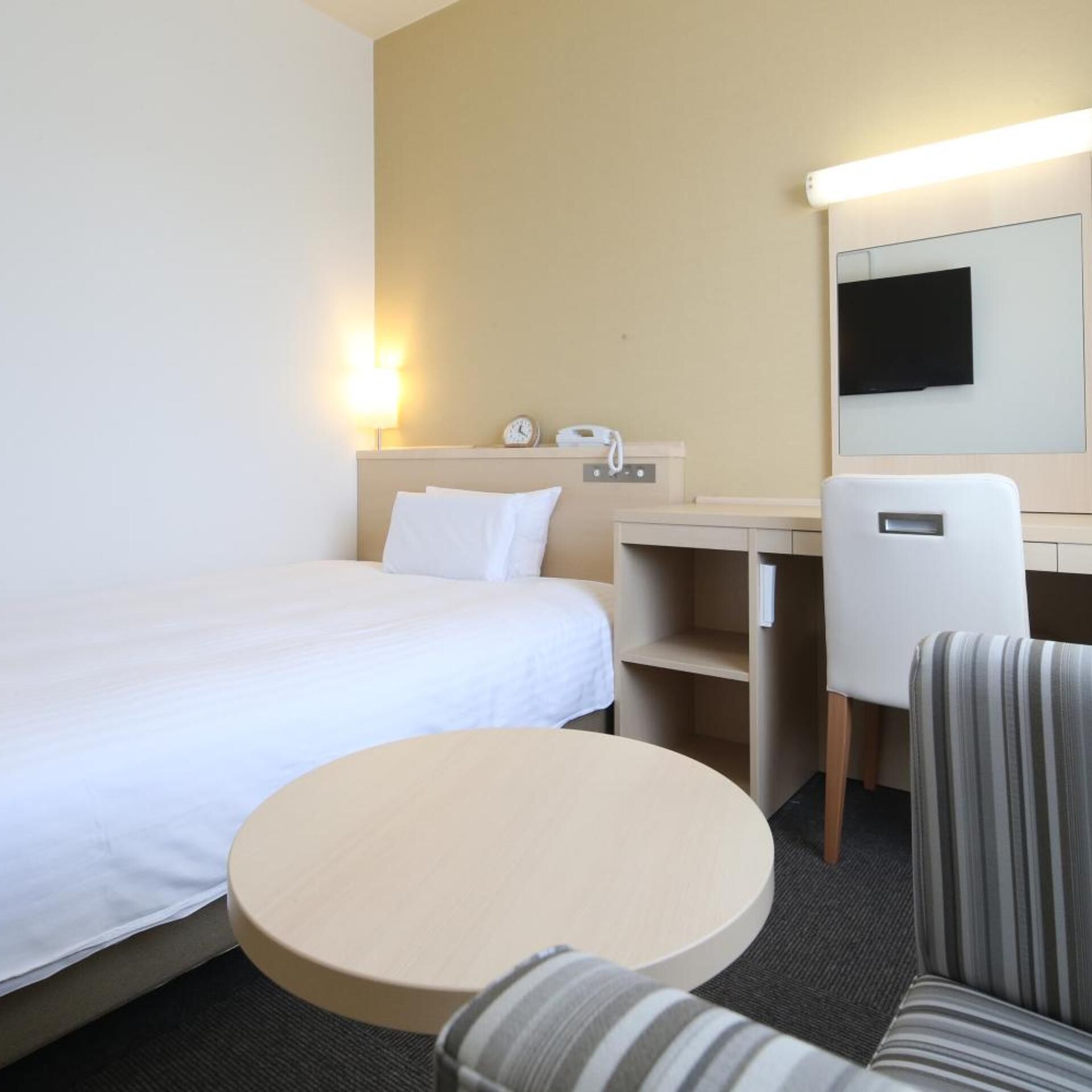 A hotel room with a bed, a desk, a chair, a round table, a wall-mounted TV, and a bedside lamp.