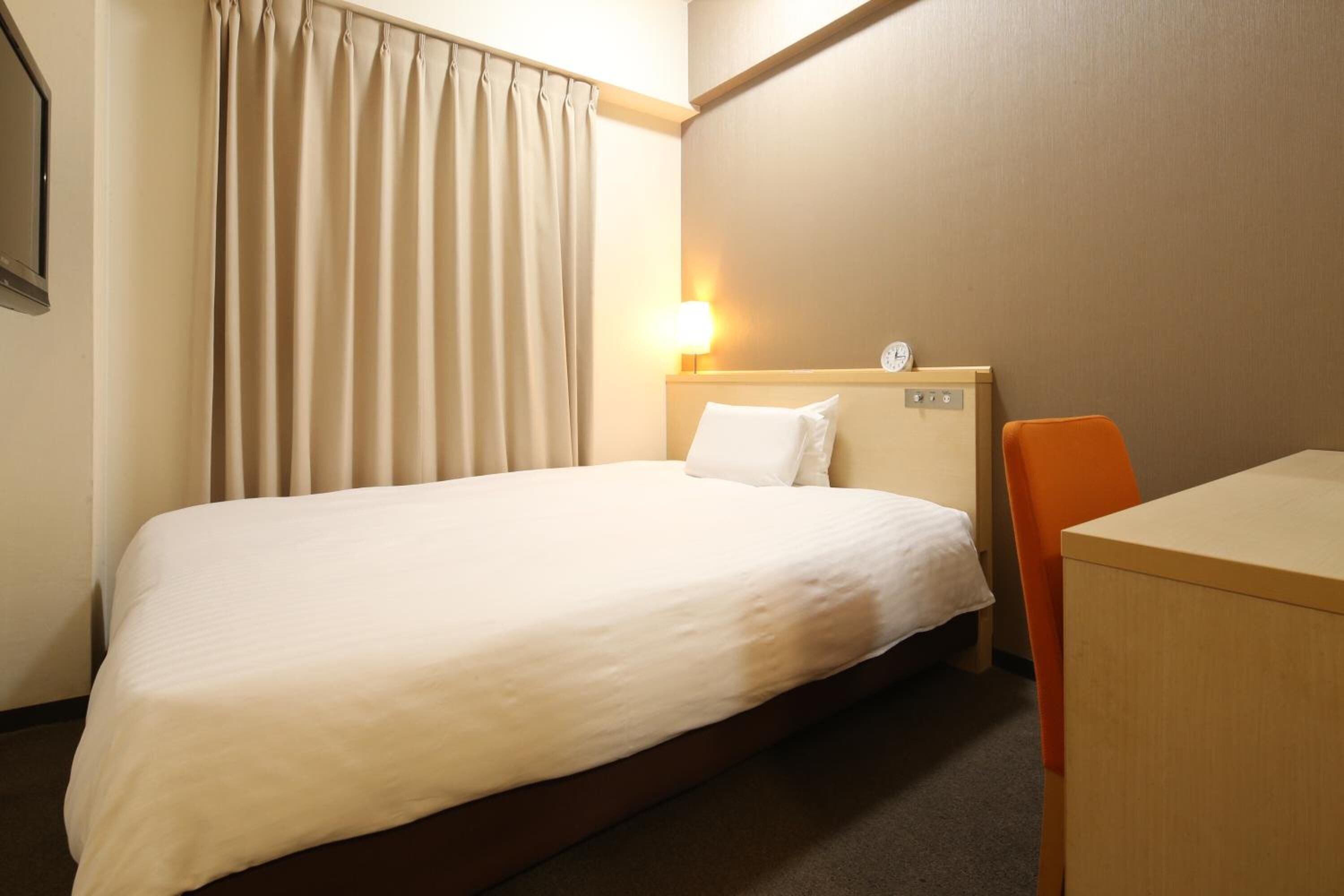 Une chambre d’hôtel avec un lit simple, un bureau, une chaise, une table de chevet avec une lampe et une horloge murale.