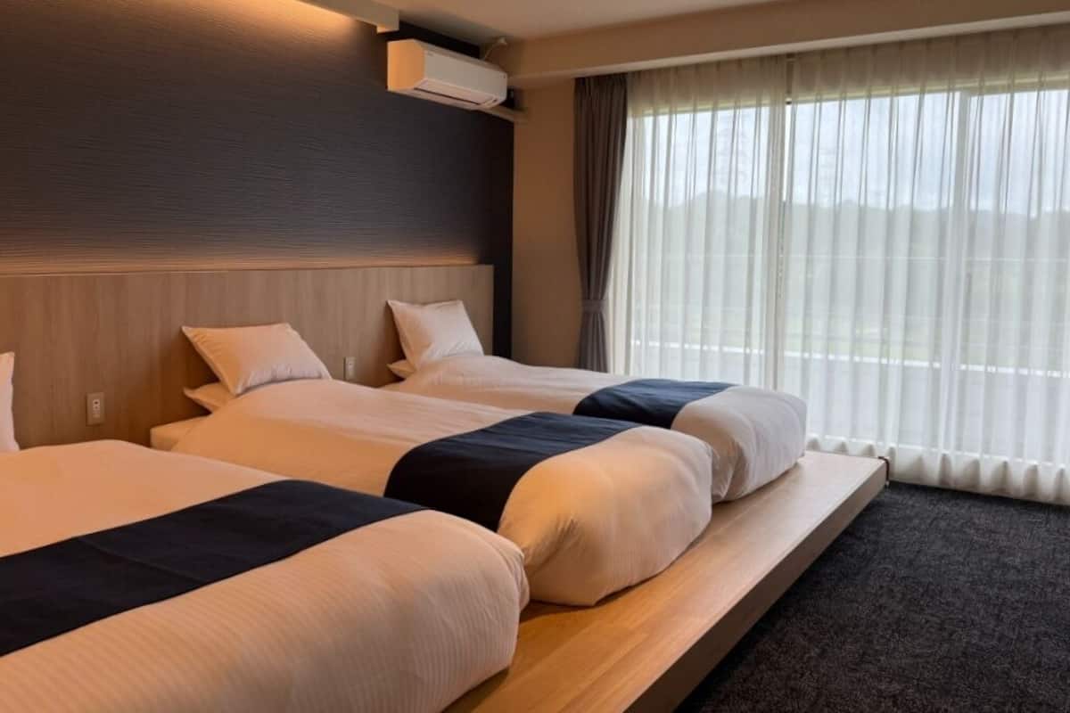 A hotel room with three beds, a wooden headboard, an air conditioning unit, and a large window with curtains.