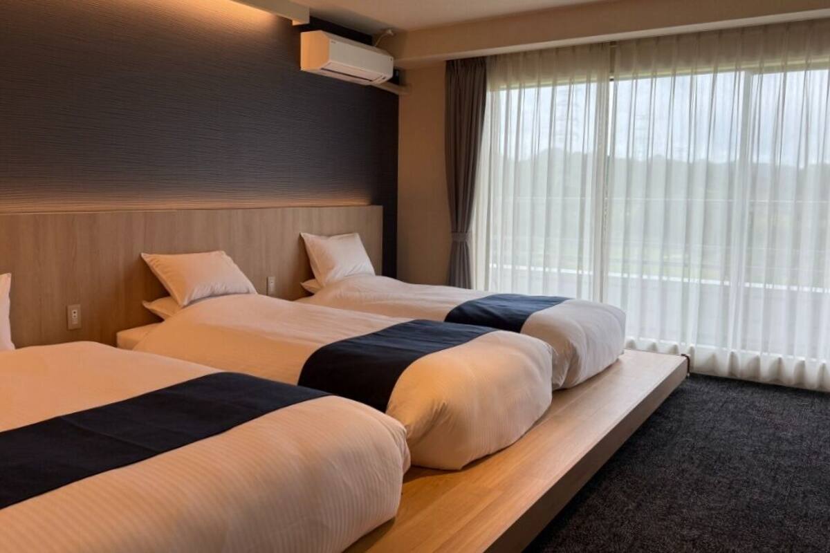 A hotel room with three beds, a headboard, an air conditioning unit, and a window with curtains.