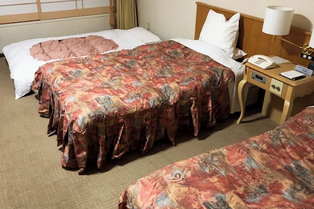 A hotel room with two beds, a bedside table with a lamp, a phone, and a small desk with a chair.