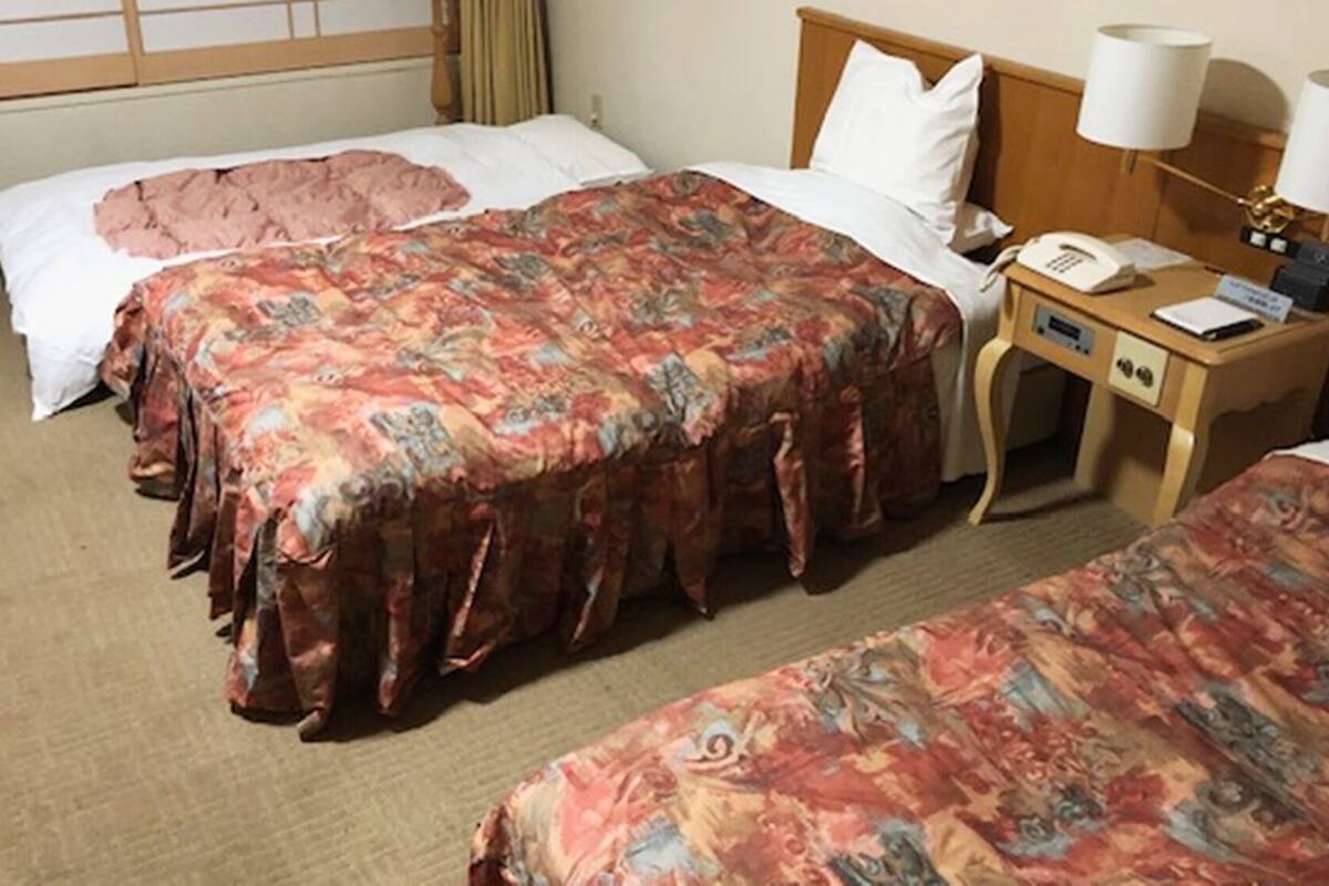 A hotel room with two beds, a bedside table with a lamp, a phone, and a small desk with a chair.