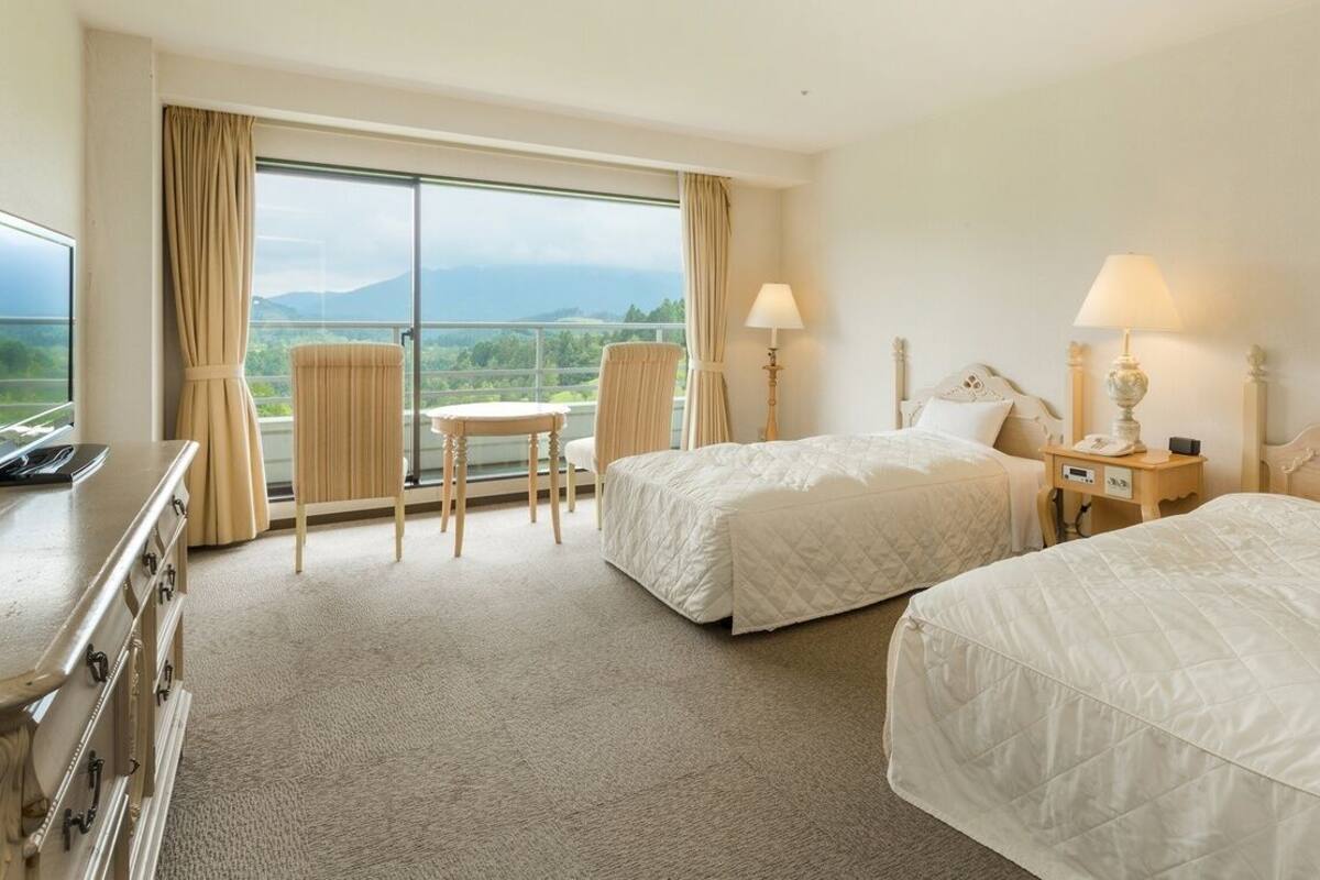 A hotel room with two beds, a desk, a chair, a table, and a view of the outdoors.