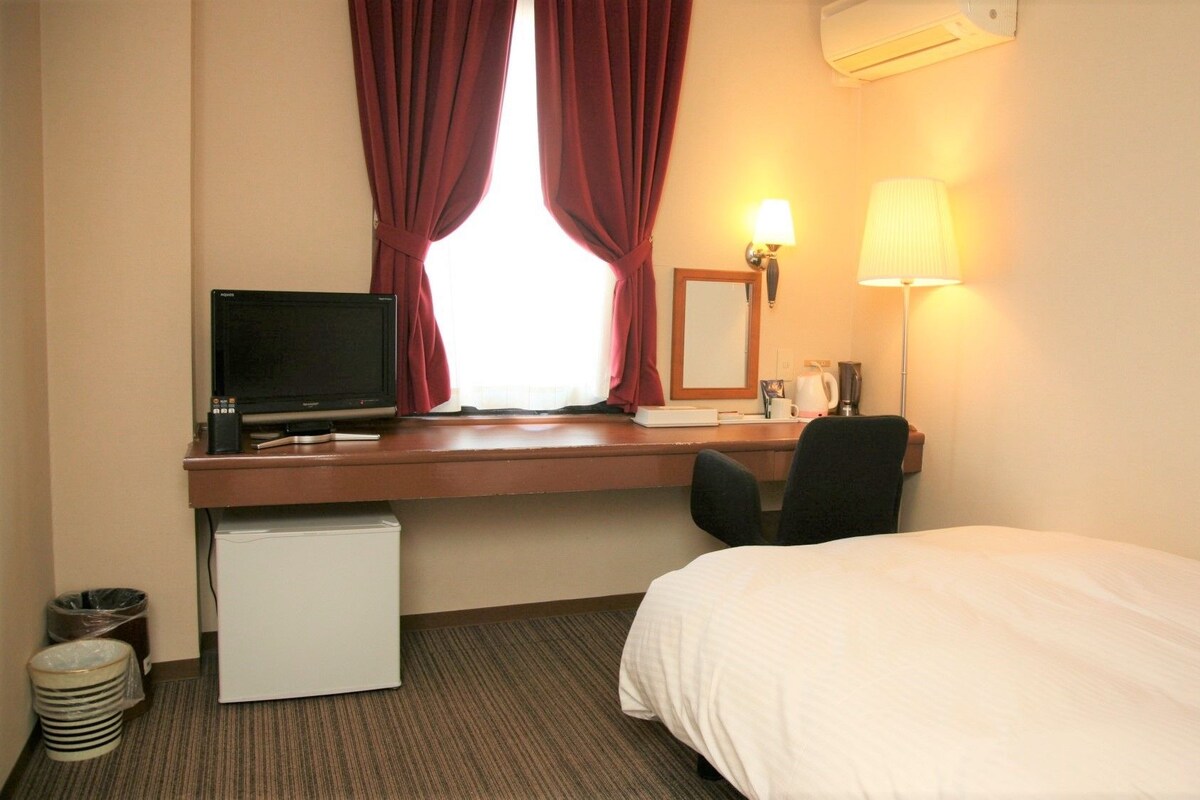 A hotel room with a bed, desk, and computer. There is a window with curtains, a lamp, and a small refrigerator.