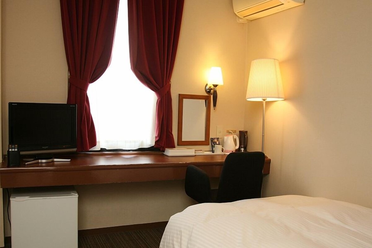 Una habitación de hotel con una cama, un escritorio con computadora, una lámpara y un refrigerador pequeño.