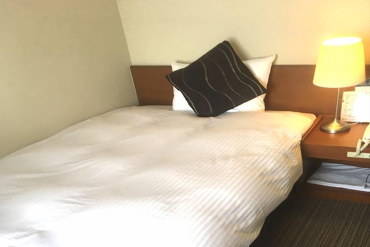 Una cama bien hecha con sábanas blancas, cabecera de color marrón oscuro y mesita de noche con lámpara y teléfono.