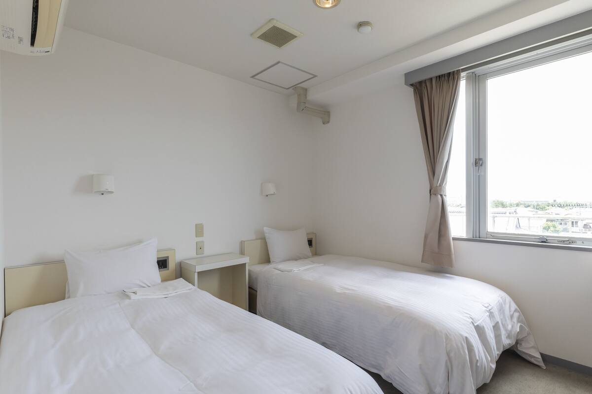 A hotel room with two beds, a window with curtains, and a view of the city.