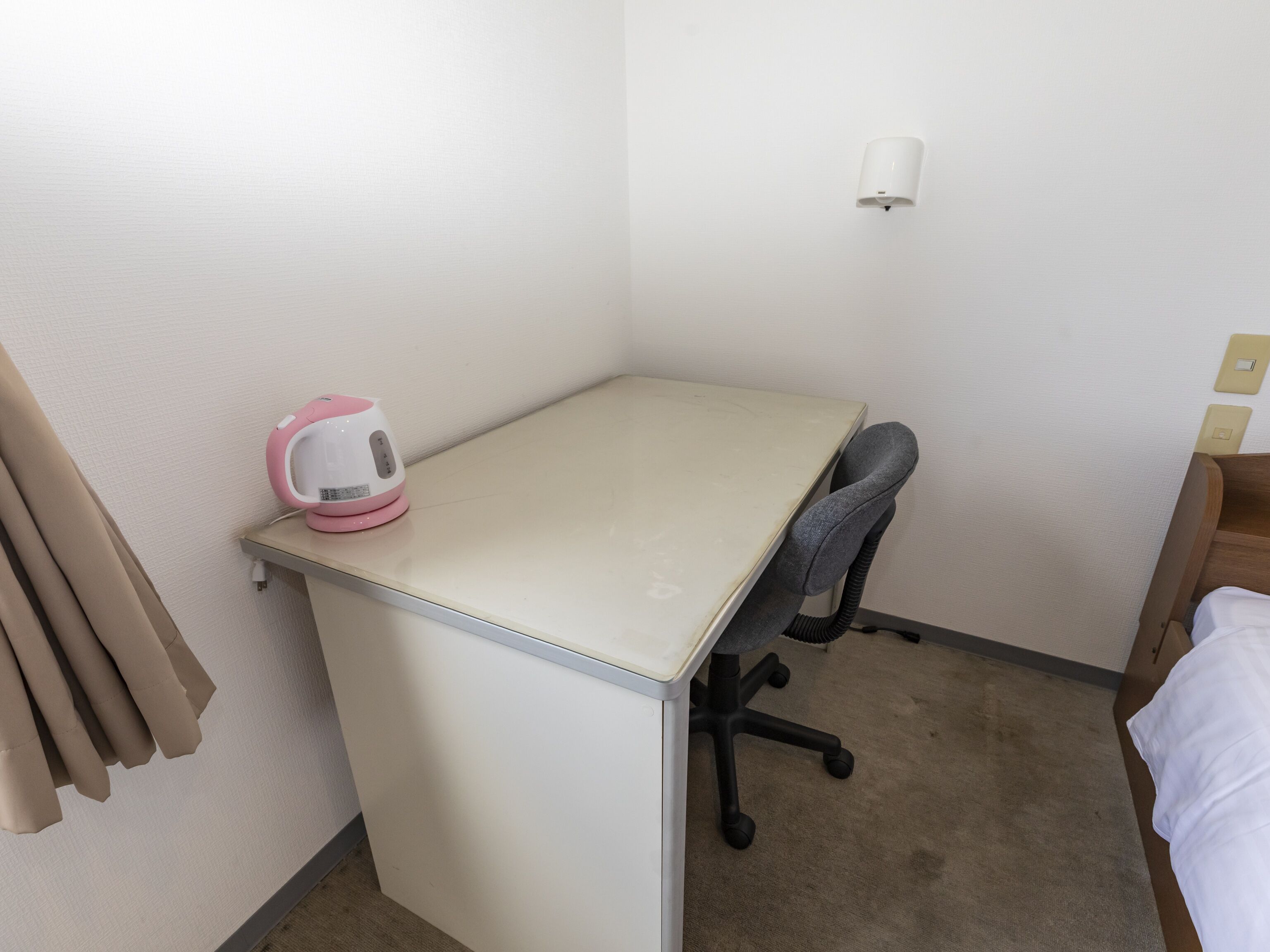 A small, single-room hotel room with a desk, a chair, a bedside table, and a bed.
