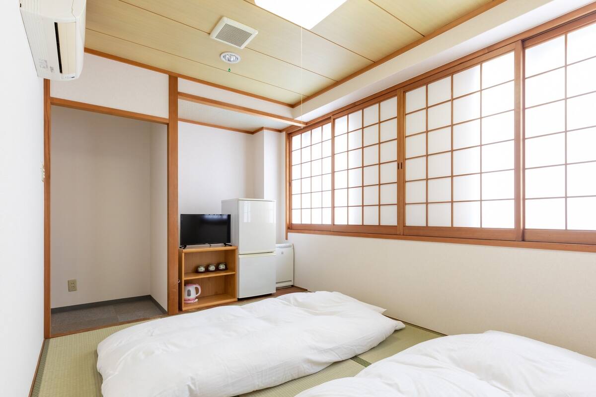 A traditional Japanese room with tatami flooring, a futon bed, a television, and a refrigerator.