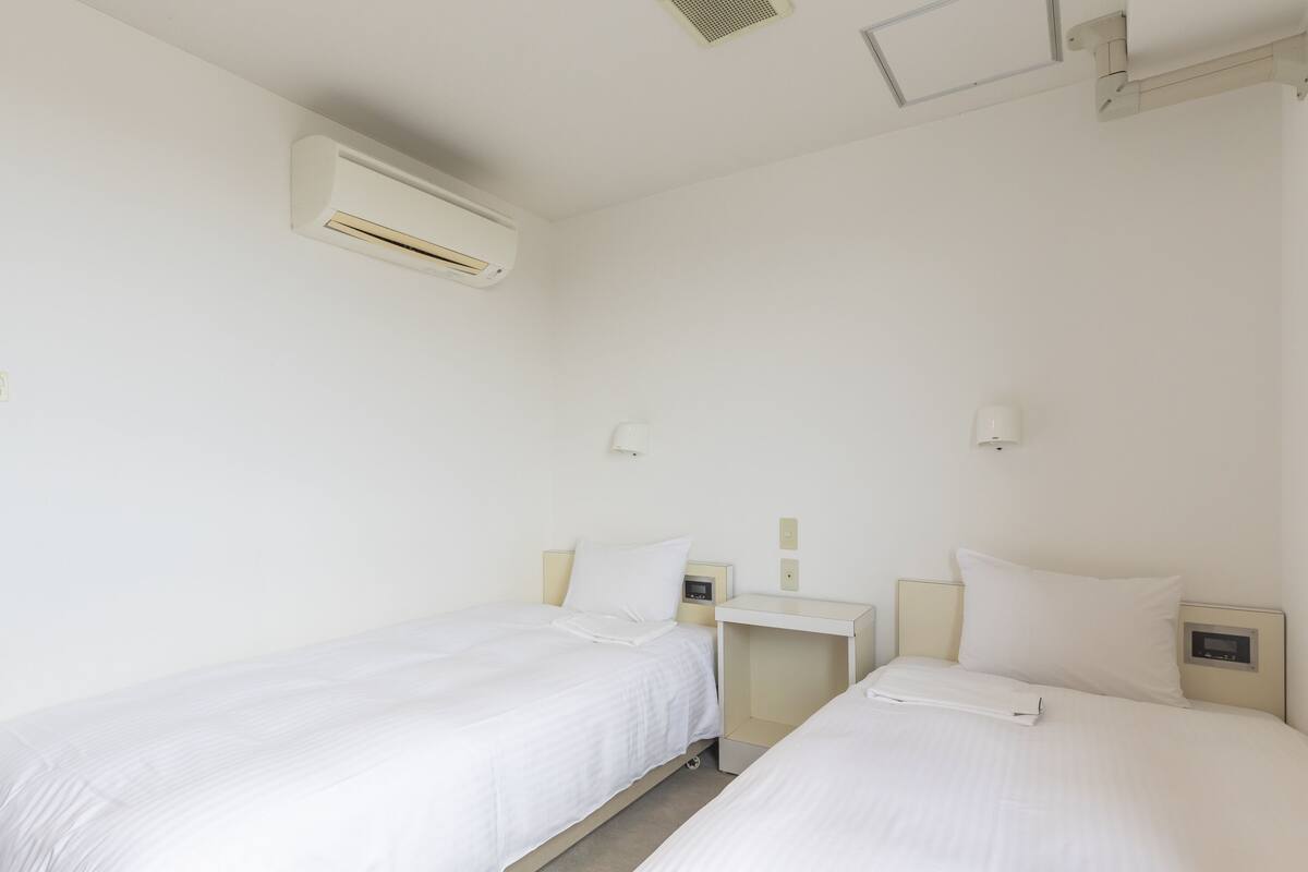 A hotel room with two single beds, a bedside table with a lamp, an air conditioning unit, and a wall-mounted light fixture.