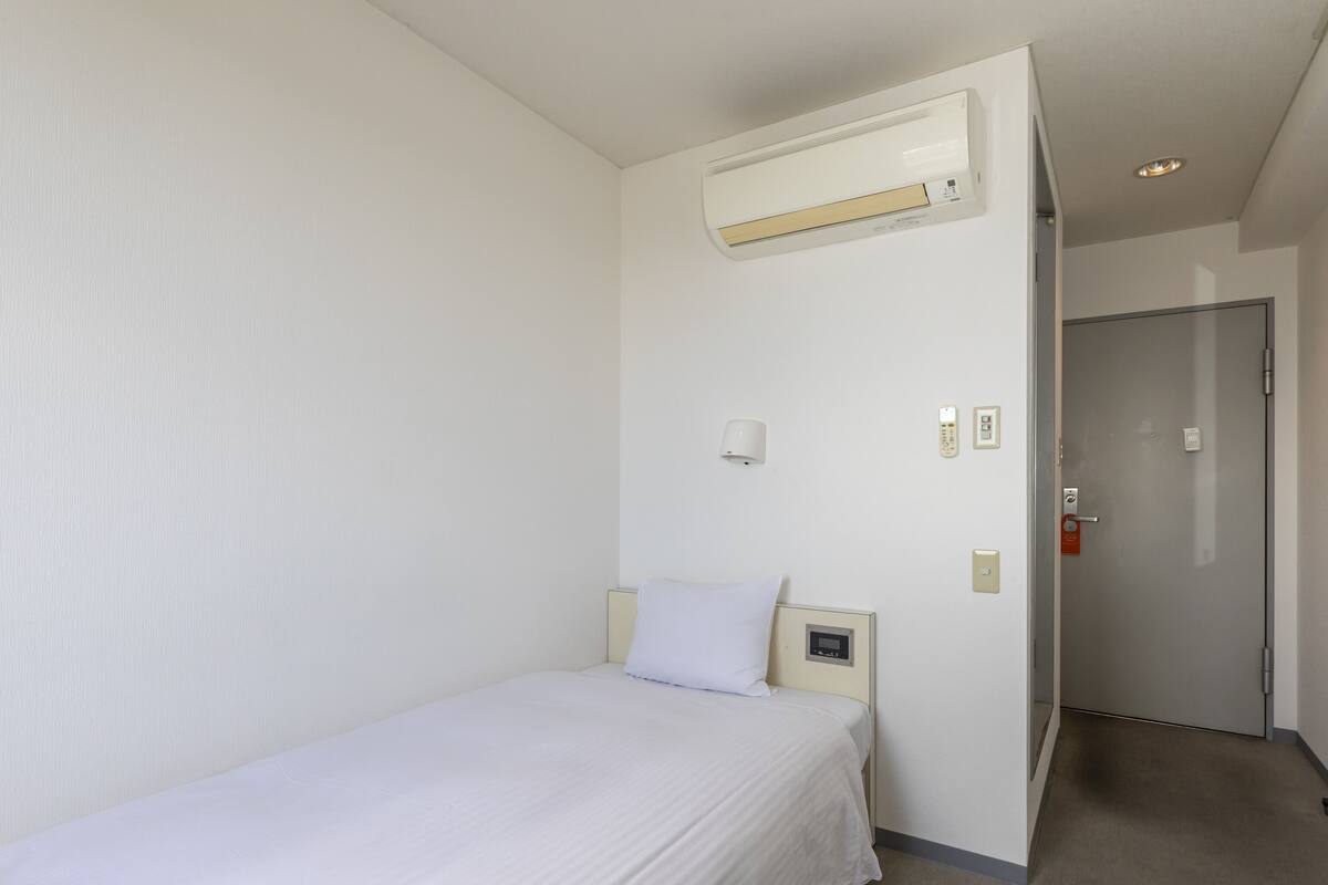 A hotel room with a single bed, a wall-mounted air conditioner, a bedside table with a lamp, and a door leading to another room.