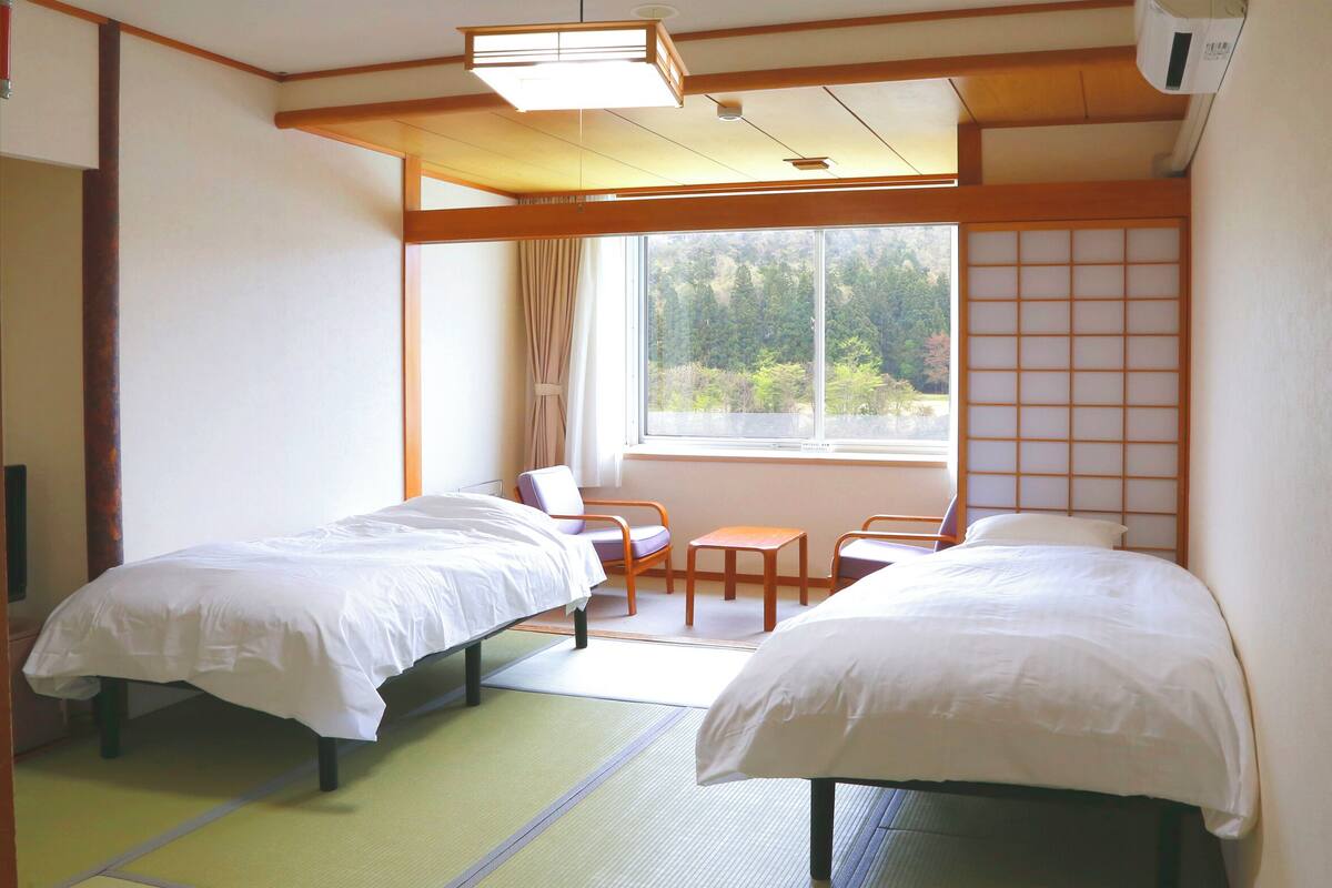 A hotel room with two beds, a window with a view of trees, a chair, and a small table.