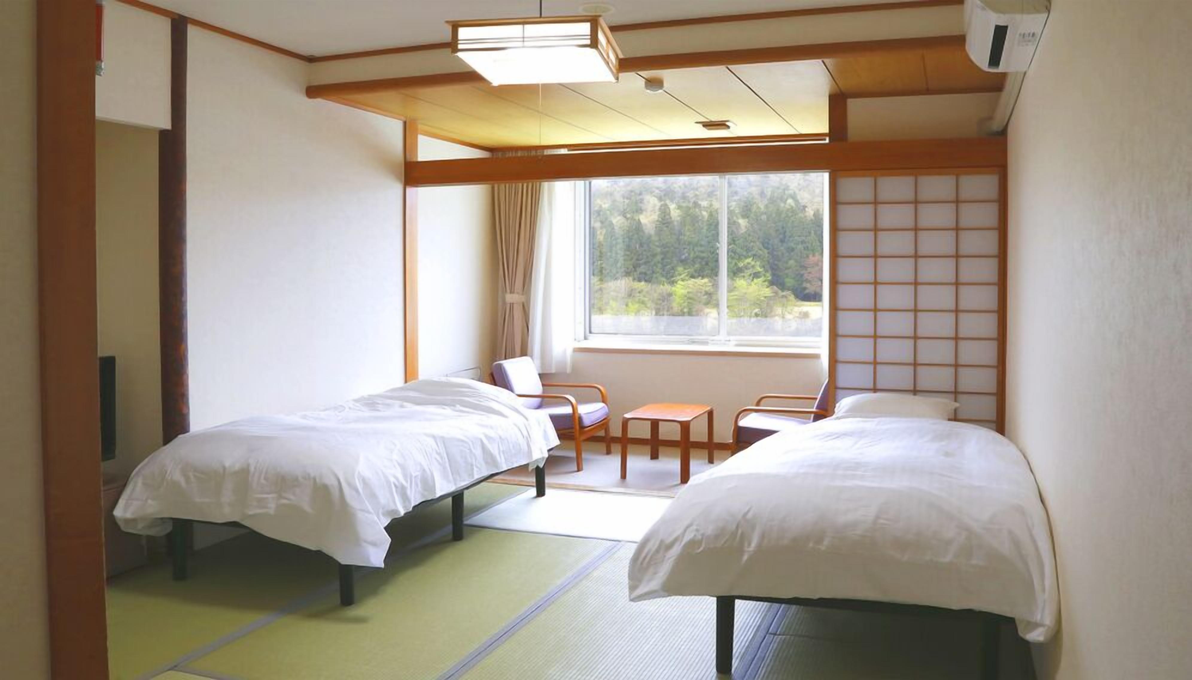 Ein Hotelzimmer mit zwei Betten, einem Fenster mit Blick auf Grünflächen, einer traditionellen japanischen Schiebetür und einem kleinen Tisch mit Stühlen.