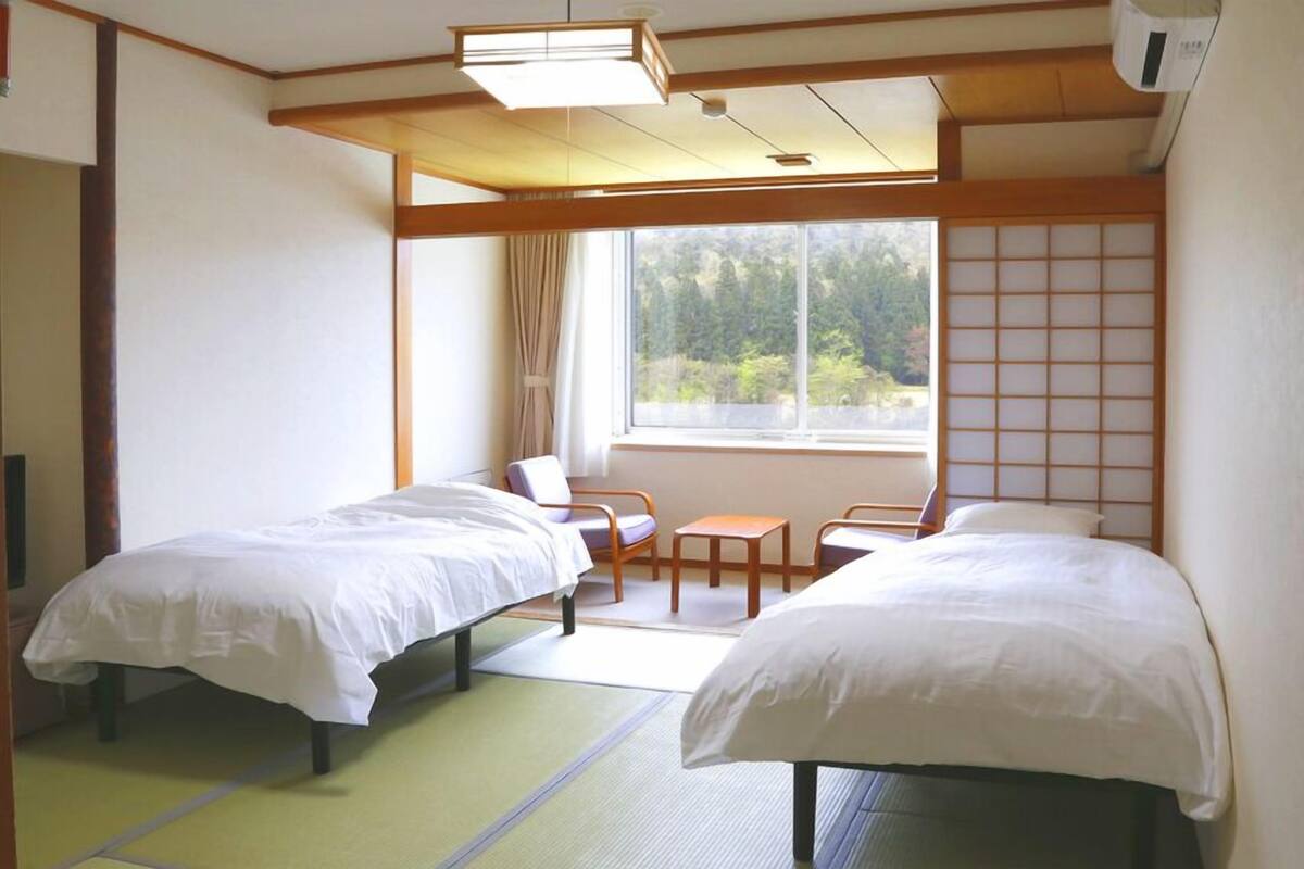 A hotel room with two beds, a window with a view of greenery, a traditional Japanese sliding door, and a small table with chairs.