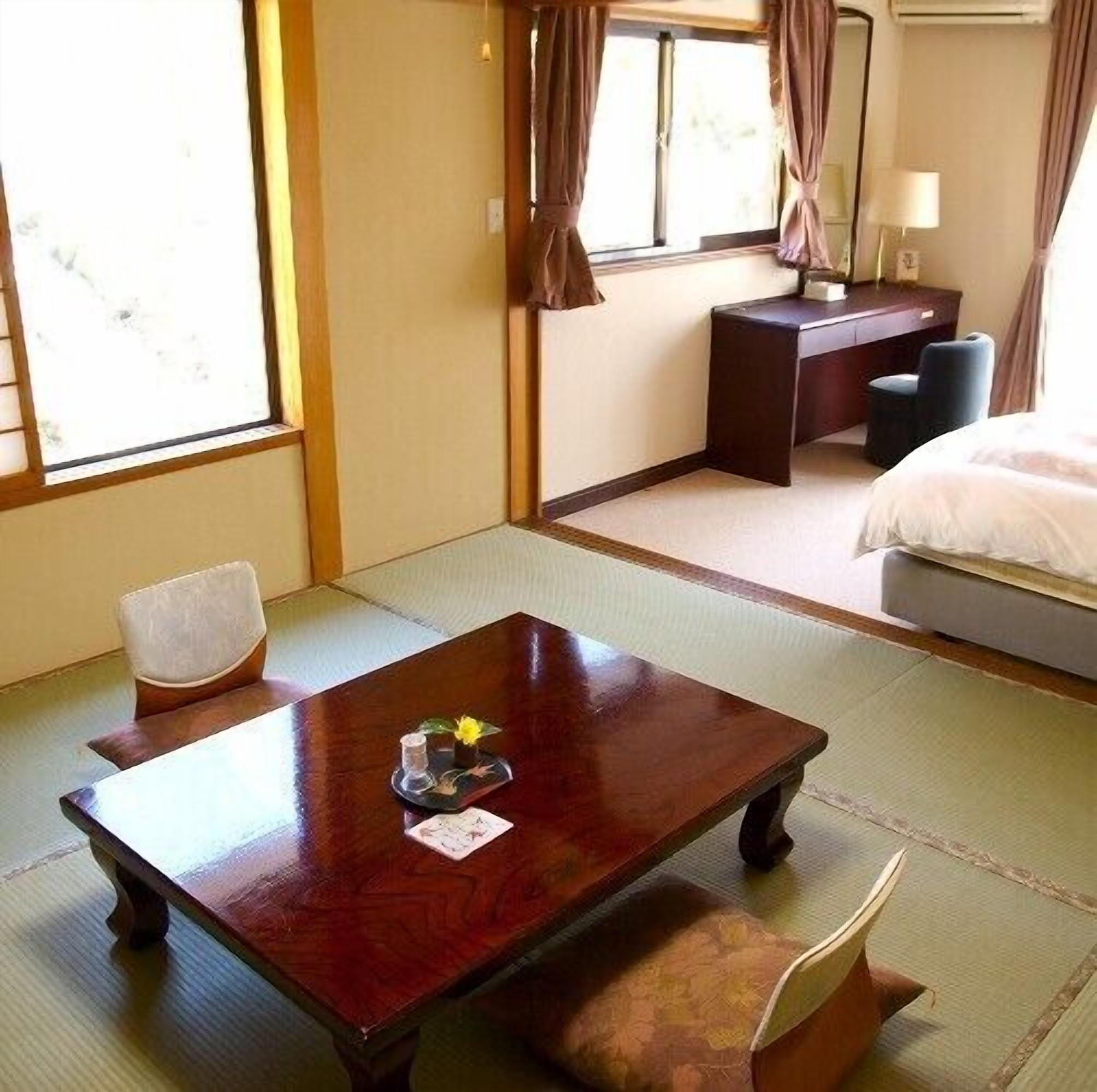 Um quarto em estilo japonês tradicional com tatames, uma mesa de madeira baixa com um conjunto de chá, uma cama com edredom branco e uma pequena escrivaninha com luminária.