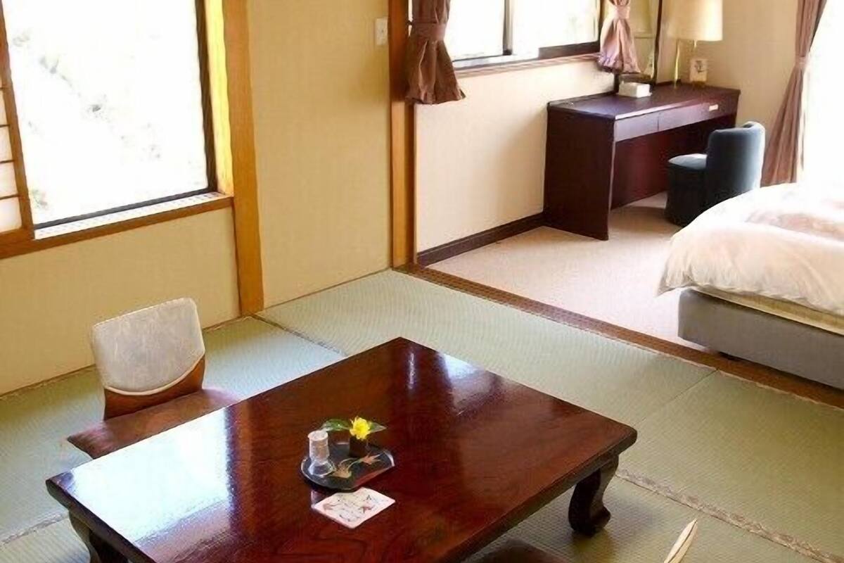 Una habitación tradicional japonesa con suelo de tatami, una mesa de madera baja con un juego de té, una cama con edredón blanco y un escritorio pequeño con una lámpara.