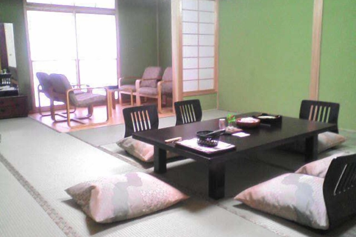 Tatami-mattoilla päällystetty huone, jossa on matala pöytä, tuoleja ja näkymä perinteiseen japanilaiseen huoneeseen.