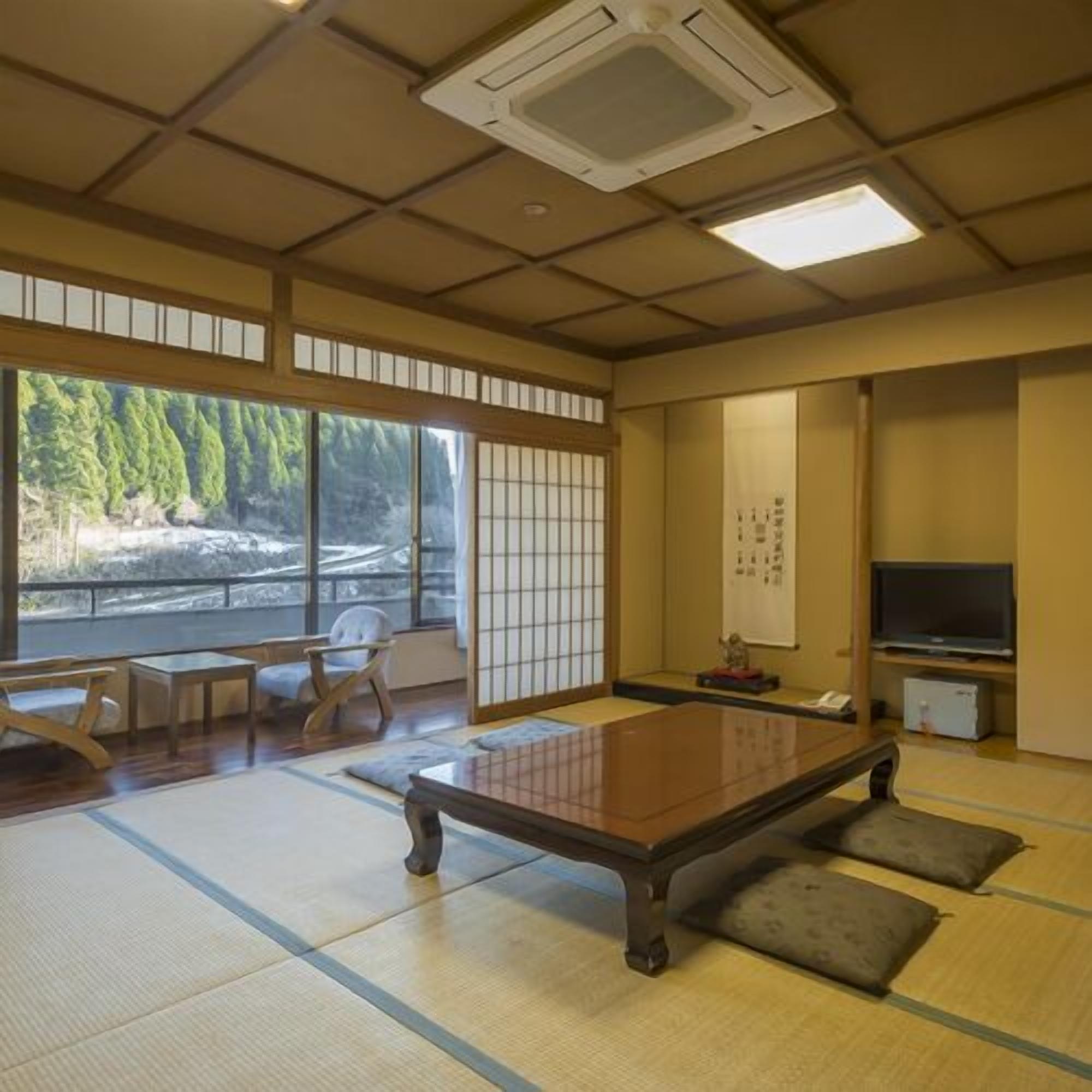 Une pièce avec un sol recouvert de tatamis, une table en tatami, une télévision et une vue sur une forêt.