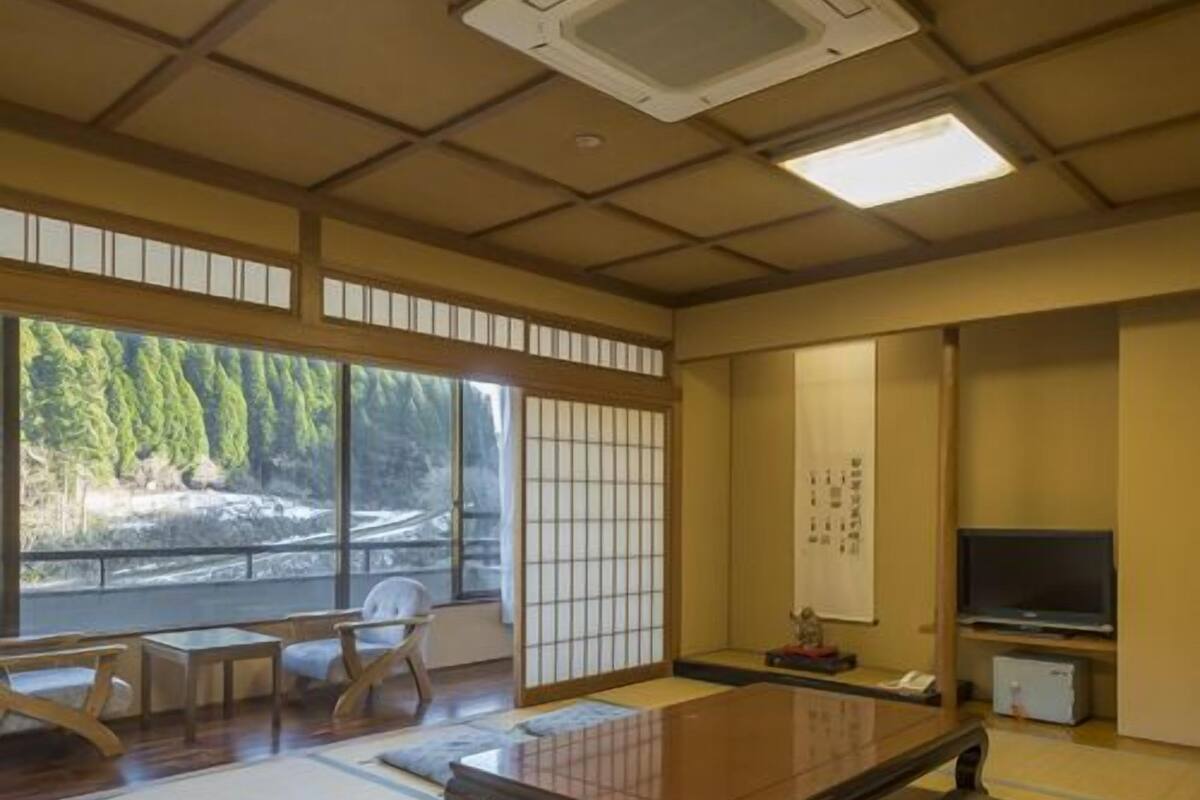 Una habitación con esteras de tatami, una mesa de tatami, una televisión y vistas a un bosque.
