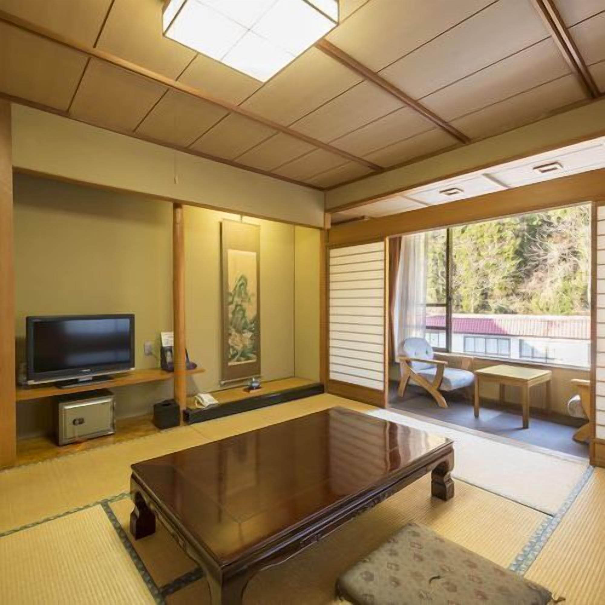 Een traditionele Japanse kamer met tatami vloerbedekking, een lage tafel, een televisie en een schuifdeur naar een buitenruimte.